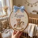 A hand holds a circular embroidery hoop featuring a personalized wooden plaque with a light blue bow, embroidered teddy bear on a rocking horse, and the name 'Lucas est. 2026' in blue script.