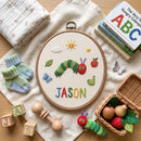 A round embroidery hoop displays a personalized cross-stitch design featuring a green caterpillar, red apple, blue butterfly, pear, sun, clouds, and the name 'JASON' in colorful letters. The hoop is styled in a flat-lay arrangement with baby accessories including a knit blanket, baby socks, wooden blocks, a rattle, a toy caterpillar, and a basket of toy vegetables on a light wood surface.