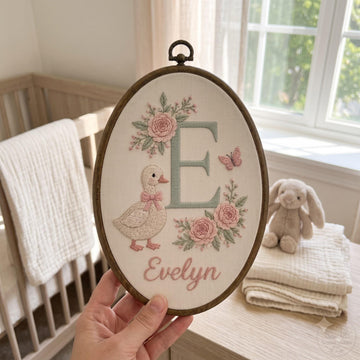 Personalized wooden embroidery hoop with a pink bow, featuring an embroidered rainbow, clouds, hearts, and the name 'Elara Fern' on a white fabric background, styled in a nursery setting.