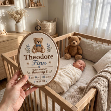 A hand holds an oval embroidered baby keepsake pillow in front of a wooden crib where a newborn baby is sleeping. The pillow features an embroidered teddy bear and personalized text reading 'Theodore Finn' with birth details.