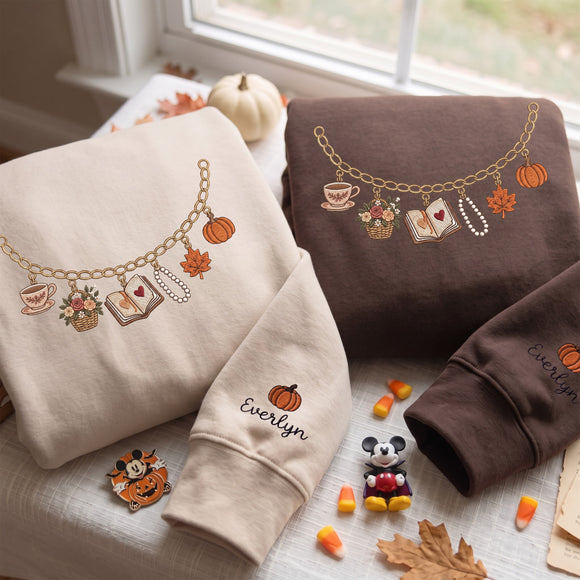 Two folded sweatshirts with embroidered autumn-themed charm necklaces and the name 'Evelyn' are displayed on a table by a window. One sweatshirt is cream and the other is dark brown, both styled with fall props like pumpkins, candy corn, and a small Mickey Mouse figure.