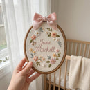 A hand holds a circular wooden embroidery hoop containing a personalized floral design with the name 'June Mitchell' stitched in the center. The embroidery features pastel pink, peach, white, and green flowers with a light pink satin bow at the top.