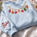 A light blue crewneck sweatshirt laid flat on a white fabric surface. The sweatshirt features a gold chain graphic across the chest with embroidered Christmas-themed charms including a gingerbread house, gift box, Santa face, Christmas tree, and reindeer.