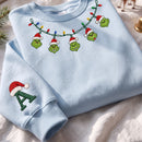 A light blue crewneck sweatshirt laid flat on a white fabric surface. The sweatshirt features embroidered Grinch-themed holiday decorations, including five green Grinch faces hanging from a string of multicolored Christmas lights and a green letter 'A' with a red Santa hat on the sleeve.