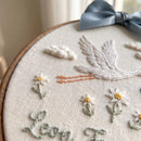 Close-up of a hand-embroidered canvas wall hanging in a wooden hoop, featuring a white stork, white daisies with yellow centers, two small white clouds, and green leaves on a cream linen background. A dusty blue satin bow is tied at the top.