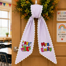 A lavender and white vertically striped personalized school ribbon with scalloped edges, hanging on a wooden classroom door. The left side of the image shows a blurred view of a classroom interior, while the right side shows a 'Welcome to Our Classroom' sign and a 'Be Kind, Be Curious, Be You!' sign.