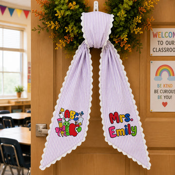 A lavender and white vertically striped personalized school ribbon with scalloped edges, hanging on a wooden classroom door. The left side of the image shows a blurred view of a classroom interior, while the right side shows a 'Welcome to Our Classroom' sign and a 'Be Kind, Be Curious, Be You!' sign.