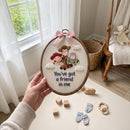 A hand holds an oval embroidery hoop featuring a custom embroidered scene of Woody, Buzz Lightyear, and Jessie with the text 'You've got a friend in me' and a pink ribbon, set against a soft nursery background with toys and a rocking chair.
