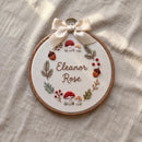 Personalized wooden embroidery hoop with a pink bow, featuring an embroidered rainbow, clouds, hearts, and the name 'Elara Fern' on a white fabric background, styled in a nursery setting.