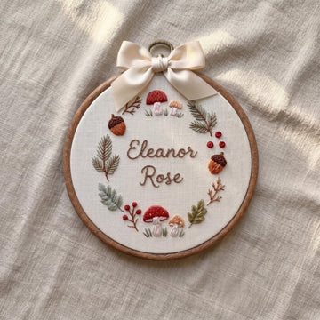 Personalized wooden embroidery hoop with a pink bow, featuring an embroidered rainbow, clouds, hearts, and the name 'Elara Fern' on a white fabric background, styled in a nursery setting.