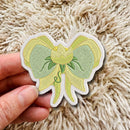 A hand holds a die-cut embroidered patch shaped like a bow and flower, featuring light green and pale yellow-green embroidery with a white stitched border, set against a fluffy cream-colored rug.