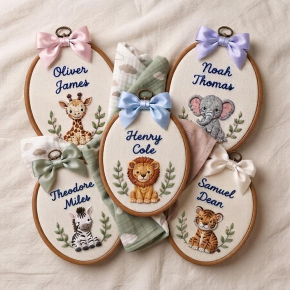 Five personalized oval embroidery hoops with safari-themed stitched animals and names, arranged on a light fabric background with a folded matching blanket. Each hoop has a ribbon bow and includes a giraffe, lion, elephant, zebra, and tiger design.