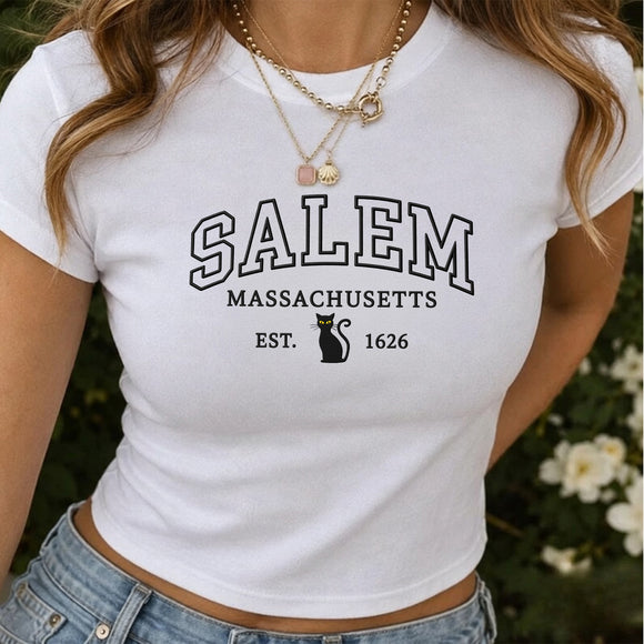 A woman wearing a white short-sleeve crop top T-shirt with 'SALEM MASSACHUSETTS EST. 1626' printed in black on the front, paired with light blue jeans and layered gold necklaces.