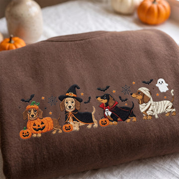 A folded brown sweatshirt with embroidered dachshunds dressed in Halloween costumes, surrounded by pumpkins and spiderwebs on a white fabric surface.