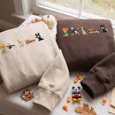 Two folded sweatshirts displayed on a table by a window: one cream and one dark brown, both featuring a horizontal embroidered line of cartoon cats in Halloween costumes. Candy corn, a small pumpkin, autumn leaves, and a Mickey Mouse charm are arranged around them.