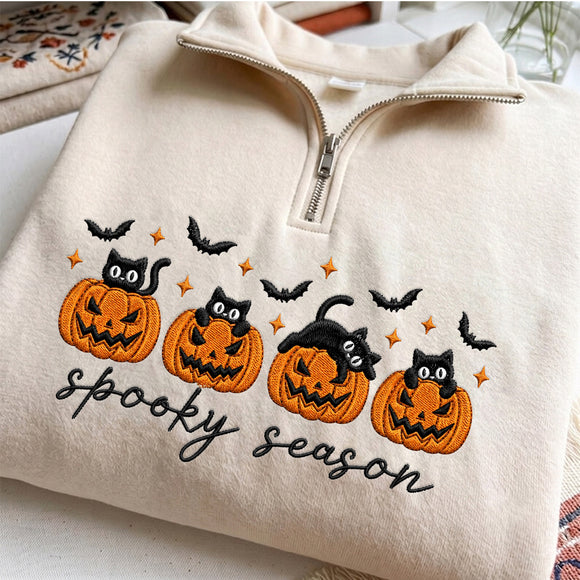 Cream-colored quarter-zip sweatshirt featuring embroidered Halloween graphics with four jack-o'-lanterns, black cats, bats, stars, and the text 'spooky season'.