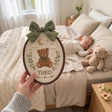 A hand holds a personalized oval embroidered patch featuring a brown teddy bear, a green bow, stars, and the name 'THEO' above a bed where a baby is sleeping next to a plush teddy bear.