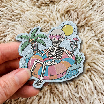 A hand holds a die-cut sticker featuring a cartoon skeleton relaxing in a beach float with palm trees, waves, and a sun, set against a beige shag rug background.