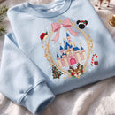A light blue crewneck sweatshirt laid flat on a white fabric background, featuring a large embroidered graphic of a fairytale castle in an oval frame with a pink bow, Christmas tree, candy cane, reindeer, and Mickey Mouse silhouette.