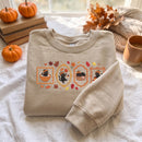 A beige crewneck sweatshirt with a fall-themed graphic of three black cats in autumn scenes, styled on a white fabric surface with pumpkins, books, and a cozy autumn background.