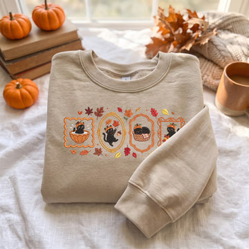 A beige crewneck sweatshirt with a fall-themed graphic of three black cats in autumn scenes, styled on a white fabric surface with pumpkins, books, and a cozy autumn background.