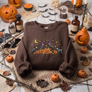 A brown crewneck sweatshirt with a colorful Halloween graphic featuring cats, pumpkins, a witch, and a crescent moon, displayed in a styled flat-lay setting with pumpkins, candles, and autumn leaves.