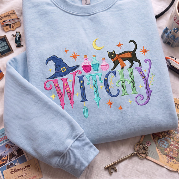 A light blue crewneck sweatshirt laid flat on a white surface, featuring a colorful 'WITCHY' graphic with a witch hat, potion bottles, a black cat, stars, and a crescent moon. Disney-themed props, including a ticket, key, and stamp, are scattered around the sweatshirt.