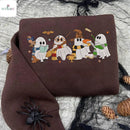 A folded brown sweatshirt with a colorful graphic of four cartoon ghosts dressed as wizards, displayed on a gray fabric background with black spiderwebs, pinecones, and a black spider decoration.