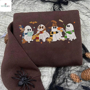 A folded brown sweatshirt with a colorful graphic of four cartoon ghosts dressed as wizards, displayed on a gray fabric background with black spiderwebs, pinecones, and a black spider decoration.